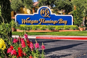 Schild für Westgate Flamingo Bay mit Logo, umgeben von Grünpflanzen und einem roten Bordstein.
