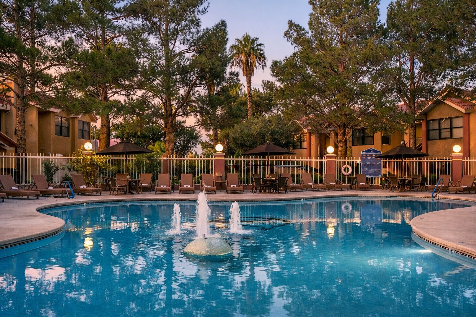 A pool with a fountain, surrounded by lounge chairs and umbrellas, with trees and buildings in the background.