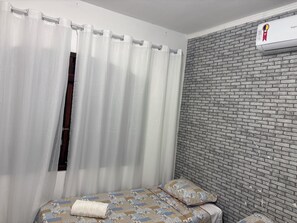 A bedroom with a bed, white curtains, a brick-patterned wall, and an air conditioning unit.