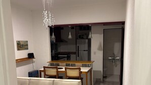 Un salon avec un canapé, une table à manger, des chaises, un lustre et, au fond, une cuisine.