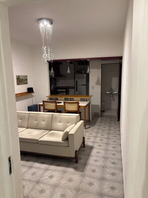 Ein Wohnzimmer mit einer Couch, einem Esstisch, Stühlen, einer Chandelüstere und einer Küche im Hintergrund.