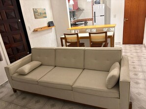 A beige sofa with cushions, a wooden dining table with chairs, and a kitchen area in the background.