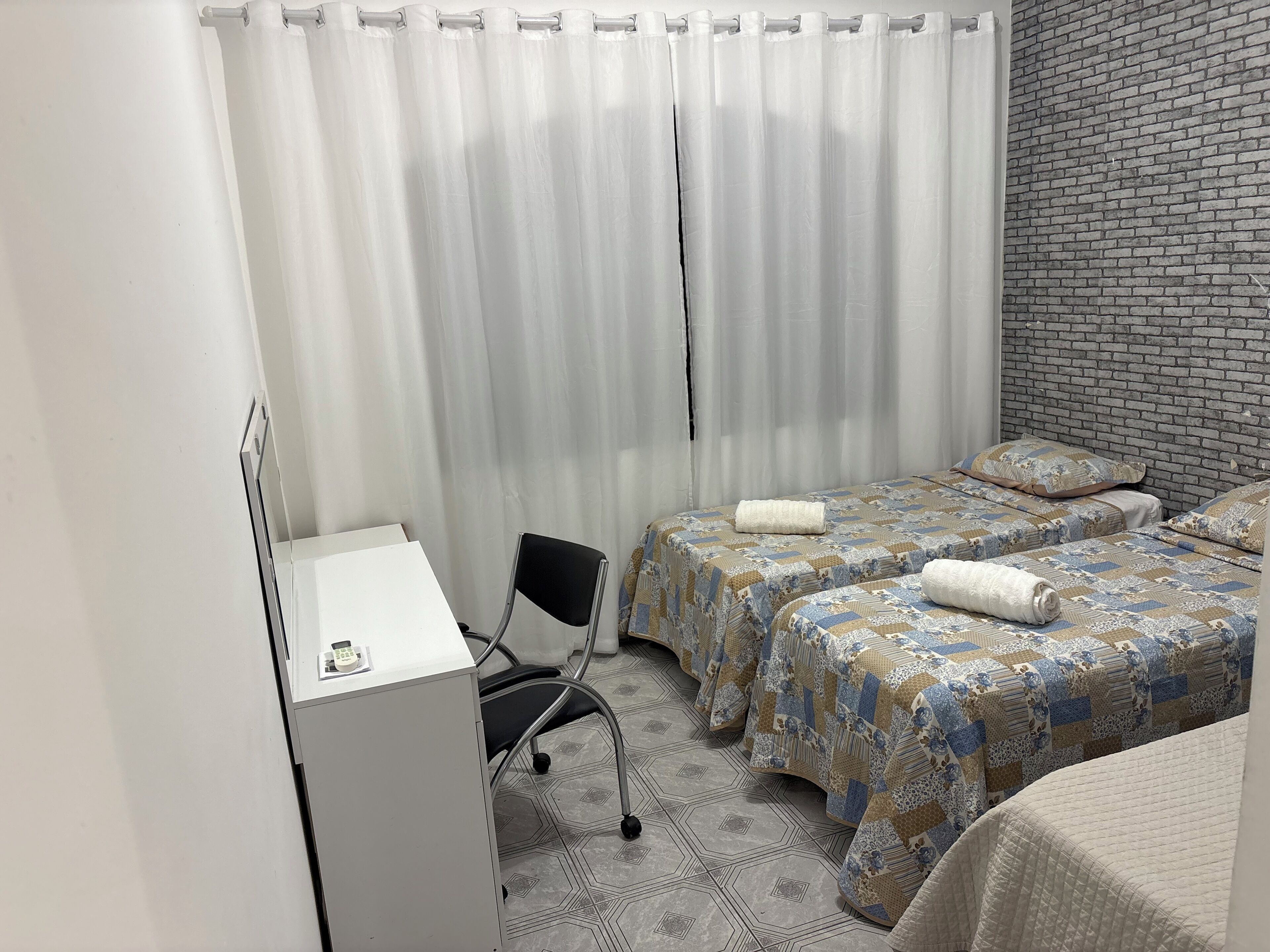A hotel room with a desk, two beds with patterned bedding, a chair, and a brick-patterned wall.