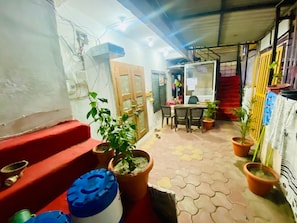 A covered outdoor area with a table and chairs, potted plants, and a red bench. - Shree Niwas Yatri Grah (Ujjain)