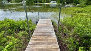 Un muelle de madera que se adentra en un lago, rodeado de vegetación y casas al fondo.