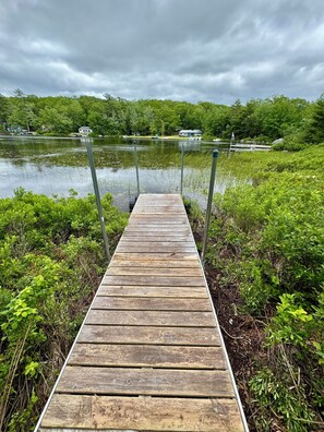 A wooden dock leading into a lake, surrounded by greenery and houses in the background.