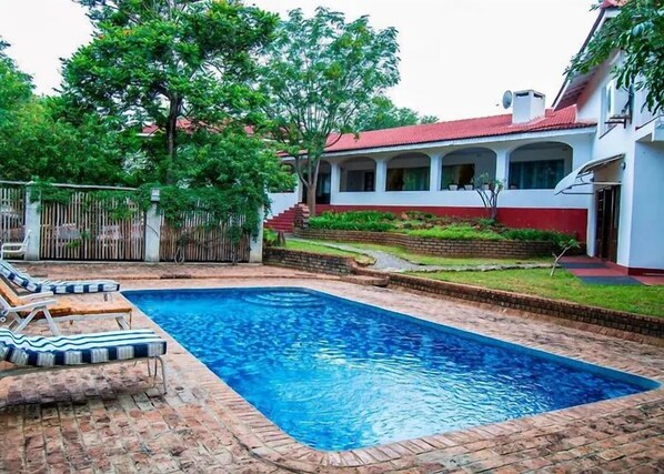 A rectangular swimming pool with a brick border, surrounded by a grassy area and a white building with a red roof.