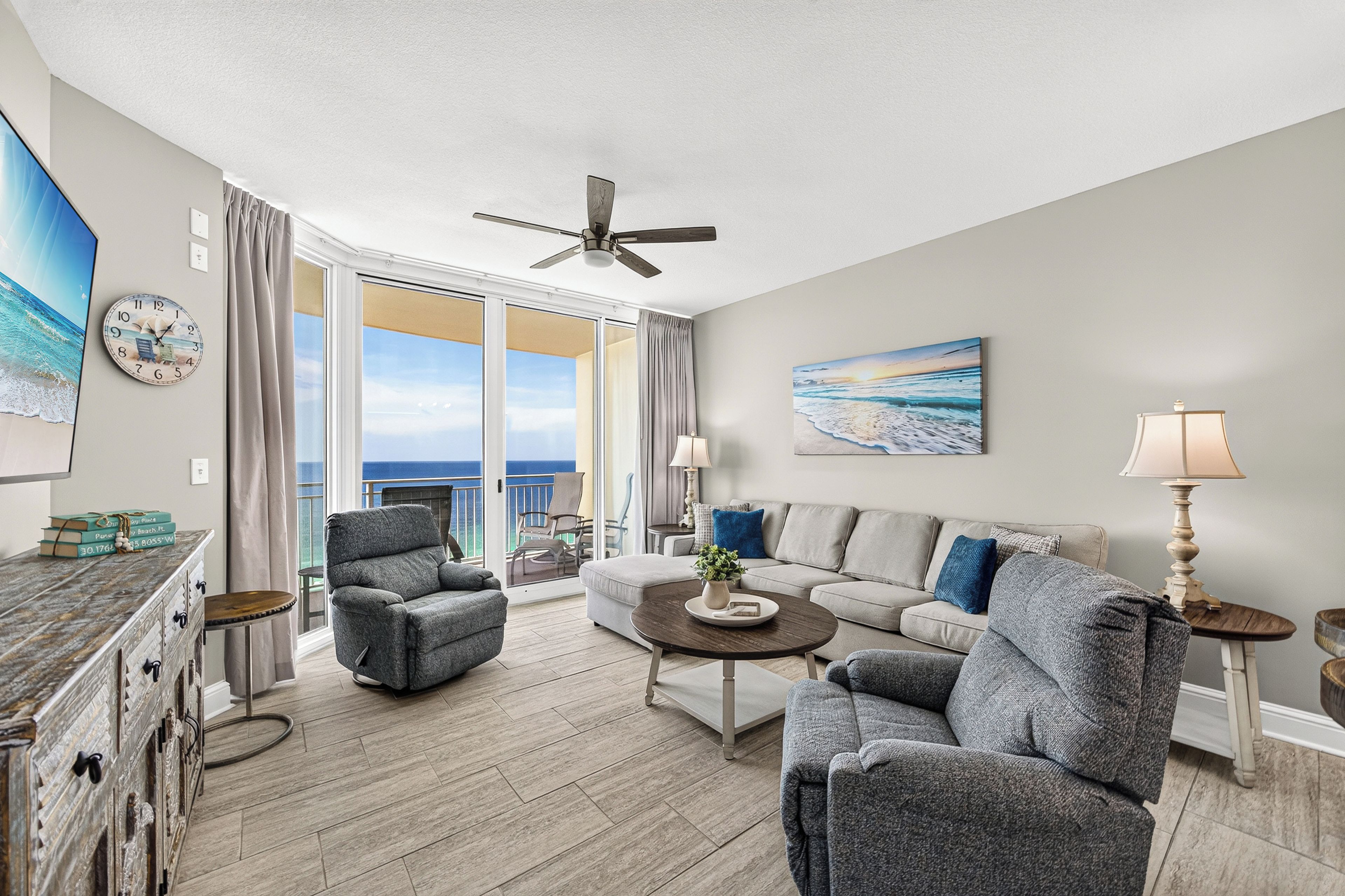 Un salon spacieux avec un balcon, un ventilateur de plafond et un tableau représentant une scène de plage.