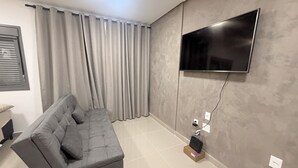 A modern living room with a grey sofa, a flat-screen TV mounted on the wall, and a window with curtains. - Modern Studio in the Best Area of ​​Mooca (São Paulo)