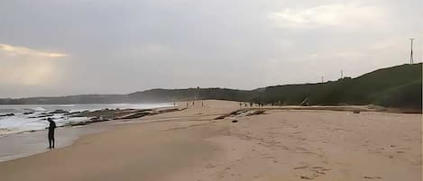 A beach with a person standing by the water, a sandy path, and distant hills.