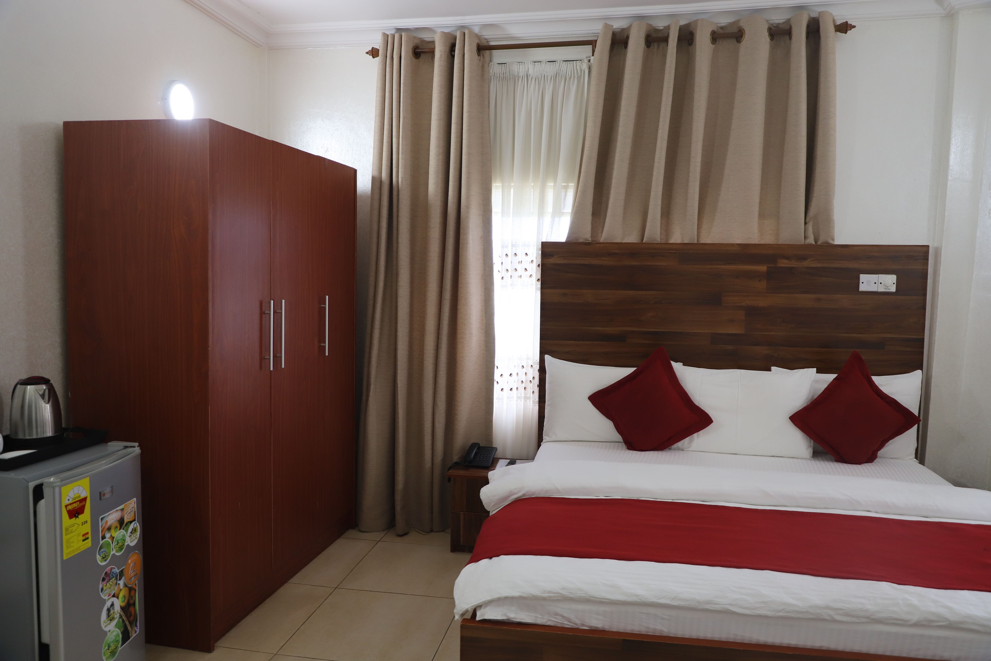 A bedroom with a wooden wardrobe, a bed with white bedding and red pillows, and a small table with a kettle.
