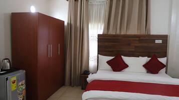 A bedroom with a wooden wardrobe, a bed with white bedding and red pillows, and a small table with a kettle.