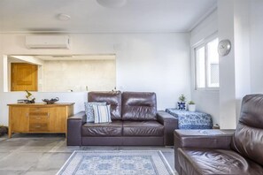 Ein Wohnzimmer mit einem braunen Ledersofa, einem Holzschrank, einem kleinen Tisch mit blau kariertem Tischläufer und einem blau gemusterten Teppich.