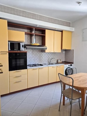 Uma cozinha compacta com armários amarelos, micro-ondas, fogão e uma área de refeições com mesa de madeira e cadeiras de metal.