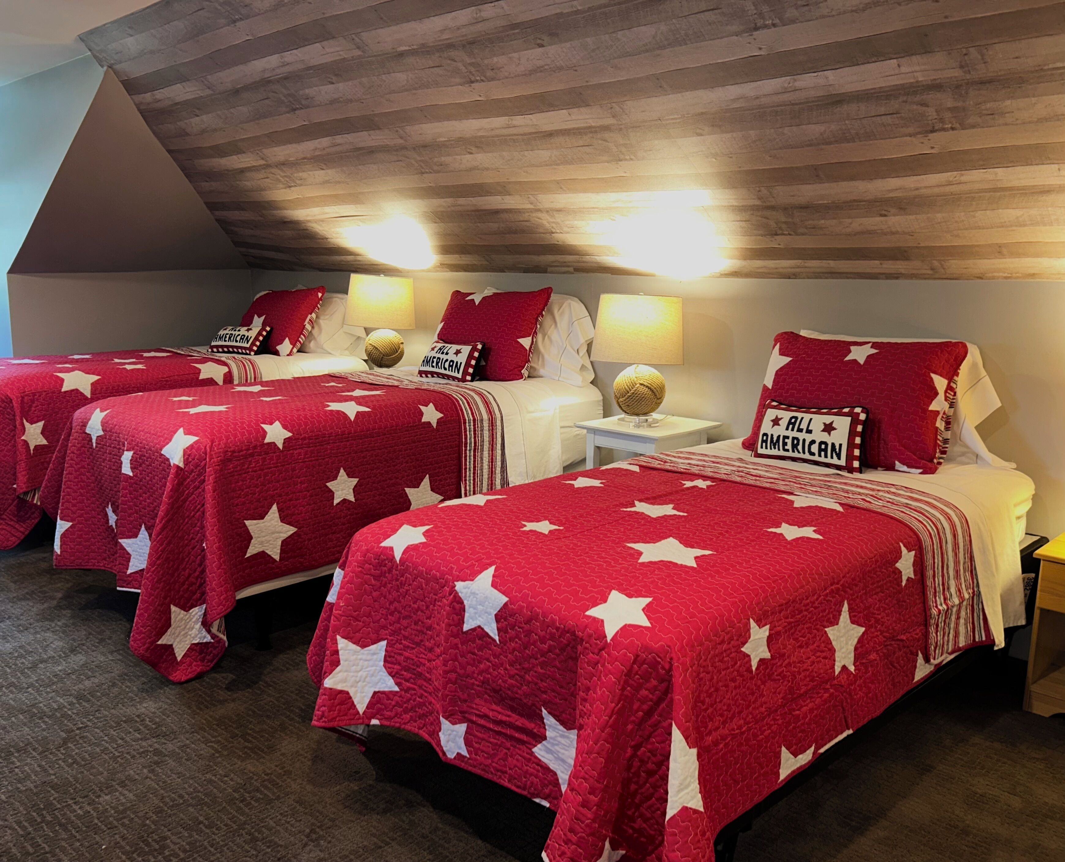 A hotel room with three beds, each with a red quilt featuring white stars and pillows with 'ALL AMERICAN' text.