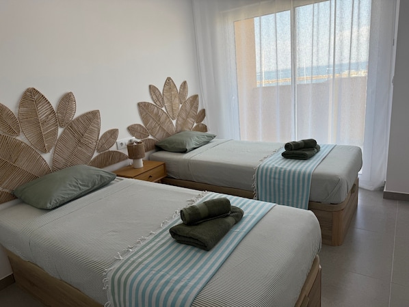 Habitación de hotel con dos camas, una mesita de noche y un ventanal con cortinas traslúcidas.