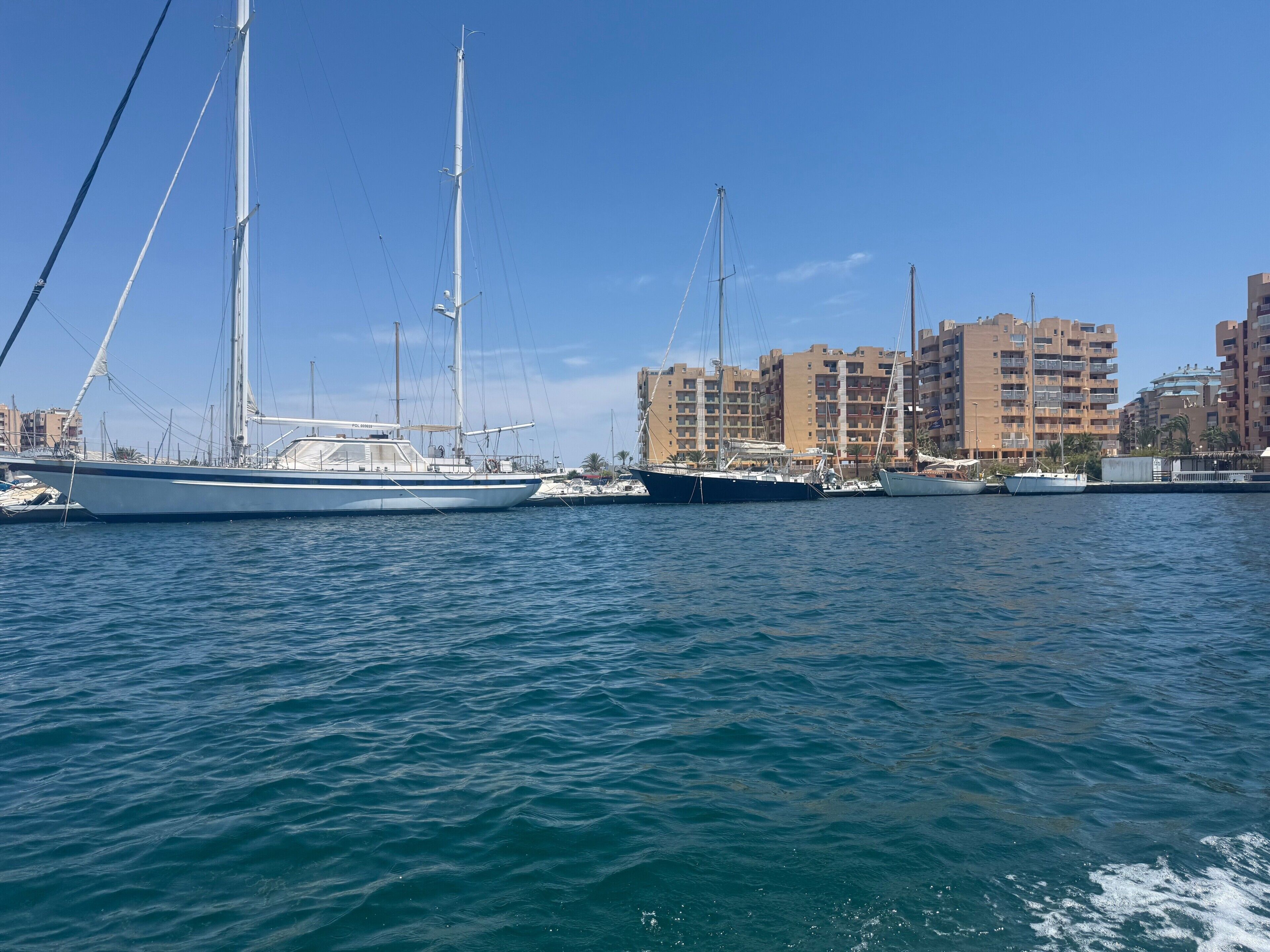 Ein Yachthafen mit mehreren Segelbooten, ein klarer blauer Himmel und Gebäude im Hintergrund.
