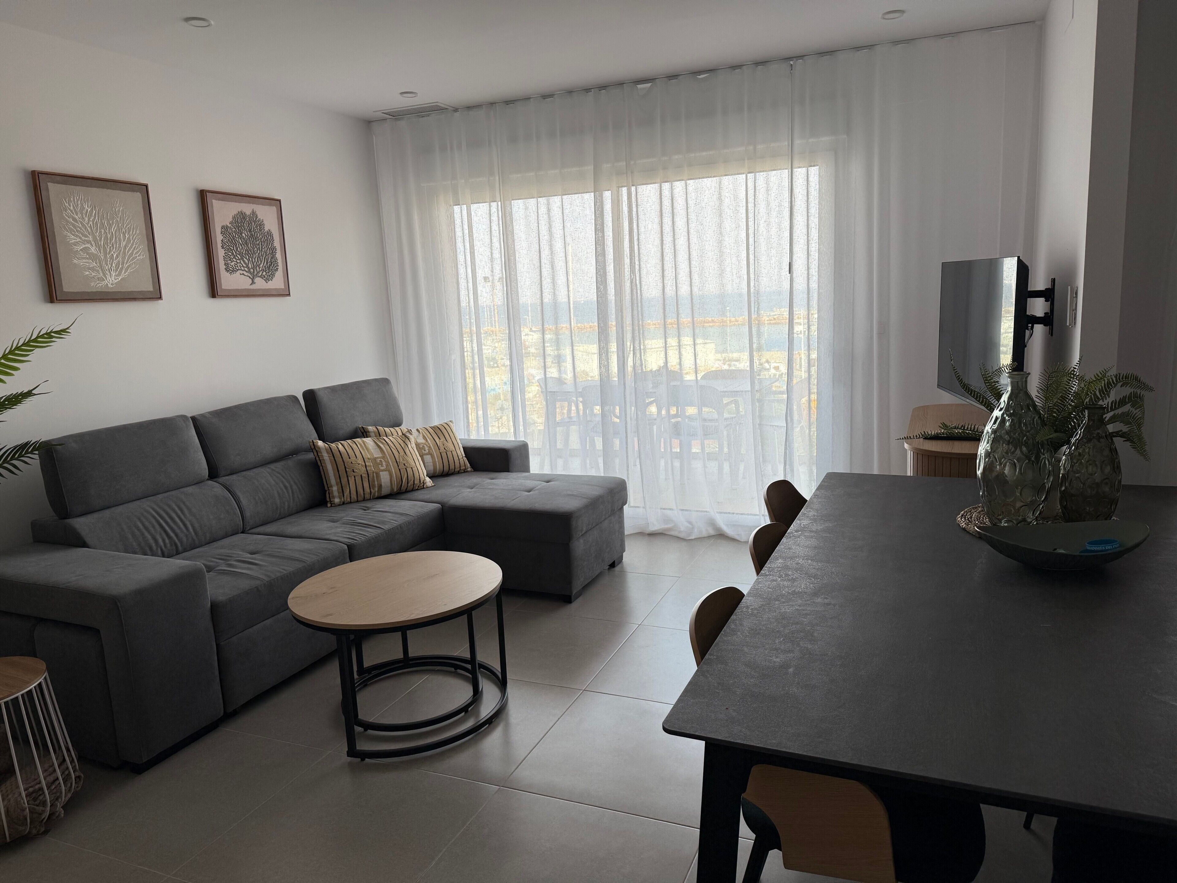 Ein modernes Wohnzimmer mit einem grauen Ecksofa, einem runden Couchtisch und einem Esstisch mit Stühlen. An der Wand hängen zwei gerahmte Pflanzenprints, und ein großes Fenster mit durchscheinenden Vorhängen.