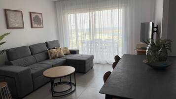 Ein modernes Wohnzimmer mit einem grauen Ecksofa, einem runden Couchtisch und einem Esstisch mit Stühlen. An der Wand hängen zwei gerahmte Pflanzenprints, und ein großes Fenster mit durchscheinenden Vorhängen.