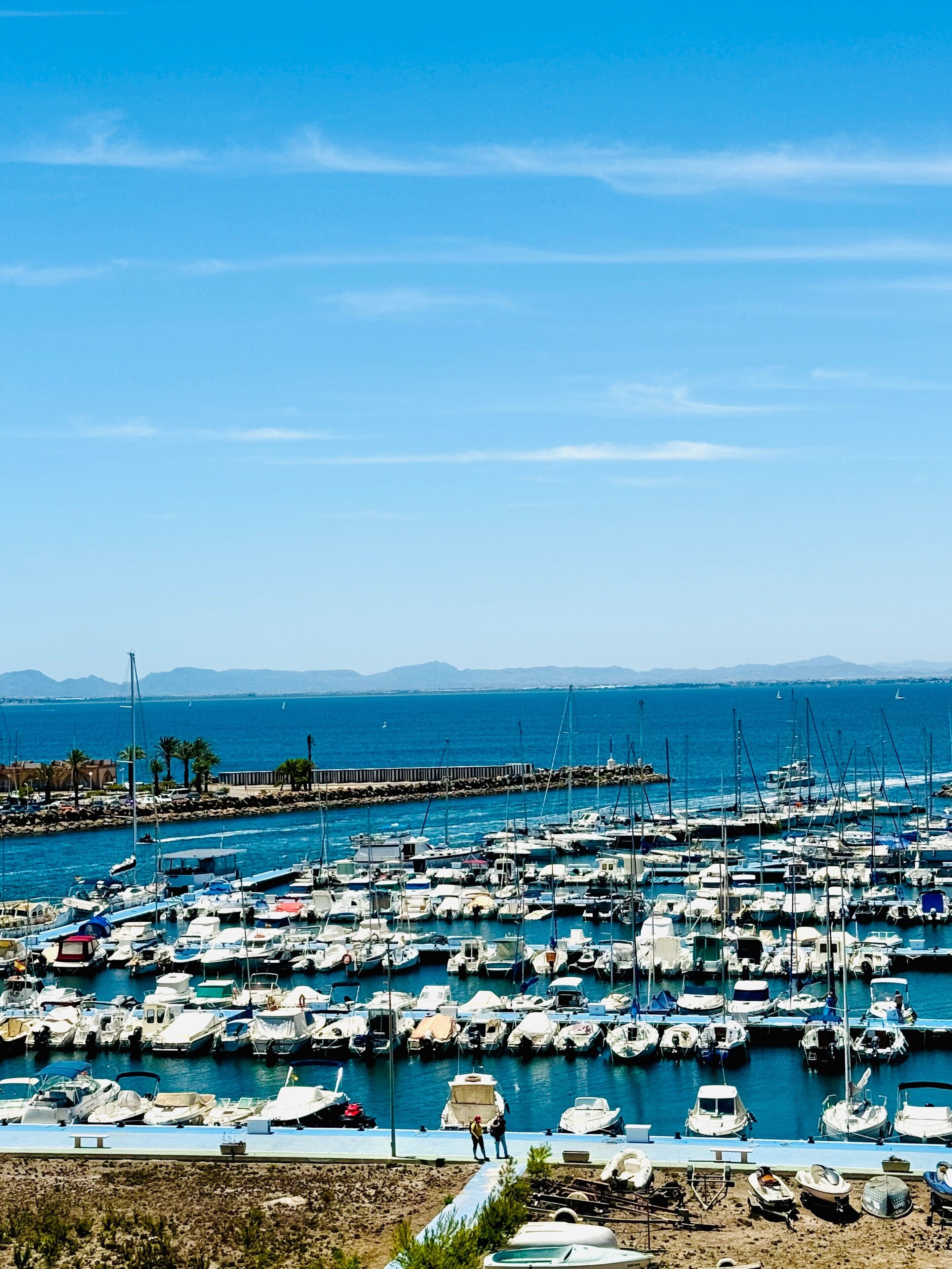 Un port de plaisance où de nombreux bateaux sont amarrés, une jetée qui s’avance dans l’eau et une vue dégagée sur les montagnes lointaines.