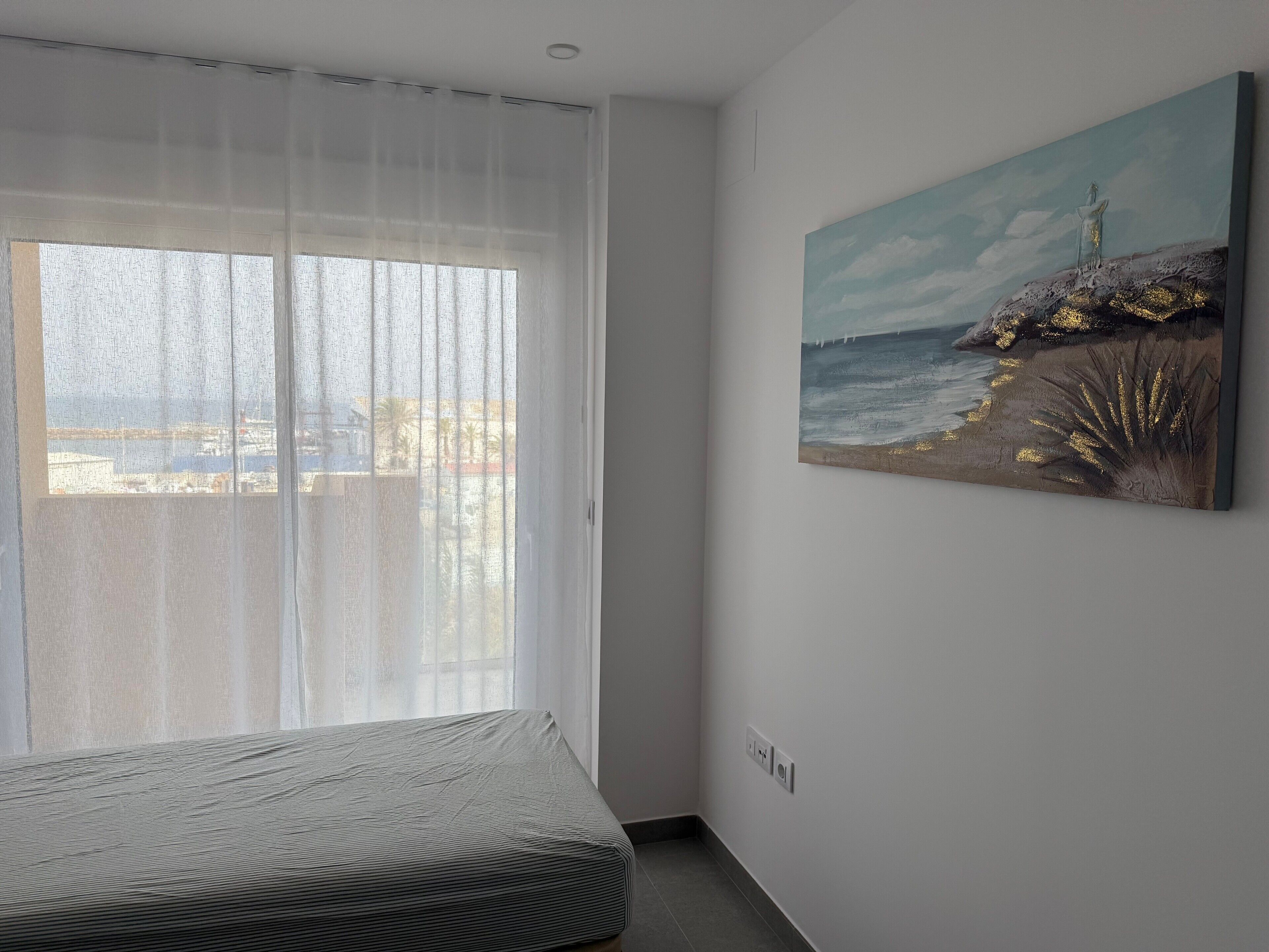 A room with a bed, a painting of a coastal scene, and a window with sheer curtains.