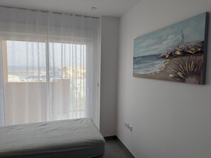 Une chambre avec un lit, un tableau représentant un paysage côtier et une fenêtre ornée de rideaux légers.