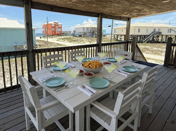 Un espace extérieur abrité, aménagé pour les repas, avec une table pour deux, donnant sur une plage et offrant une vue sur des bâtiments en arrière-plan.