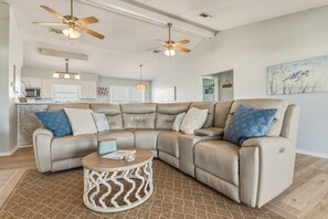 A modern living room with a sectional sofa, two ceiling fans, a round coffee table, and a kitchen in the background. - Sea Glass, Gulf Beachside, 3/2, Sleeps 9 (Dauphin Island)