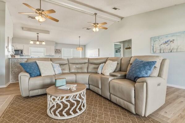 A modern living room with a sectional sofa, two ceiling fans, a round coffee table, and a kitchen in the background.