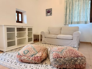 A living room with a white sofa, a white cabinet with glass doors, and two colorful poufs on a patterned rug.