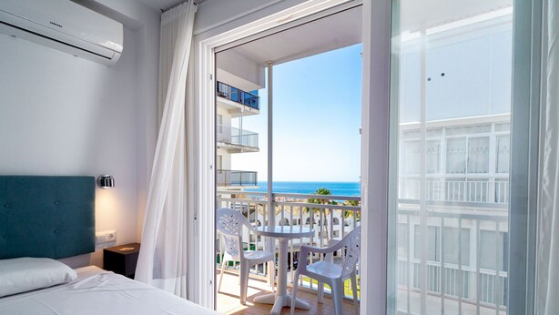 Een hotelkamer met een bed, nachtkastje, balkon met een tafel en stoelen, en uitzicht op zee.