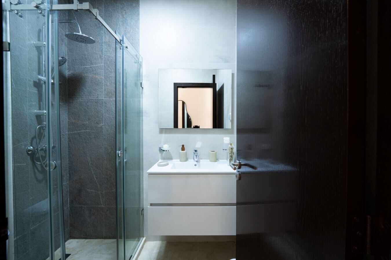 Un baño con cabina de ducha de vidrio, un lavabo blanco con espejo encima y una puerta de madera oscura.