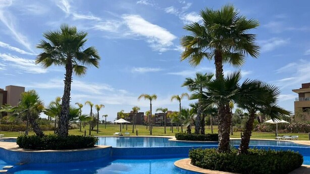 A swimming pool with palm trees, a clear sky, and buildings in the background. - Prestigia Golf Resort |Luxury 3BR| Pool&Atlas View (Marrakesh)