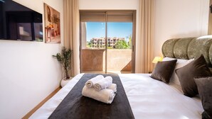 A hotel room with a bed, a TV, a balcony with a view of buildings, and a plant. - Prestigia Golf Resort |Luxury 3BR| Pool&Atlas View (Marrakesh)