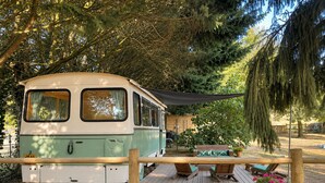 Un camping-car vintage est garé dans une zone boisée, avec une terrasse en bois et une table de pique-nique.