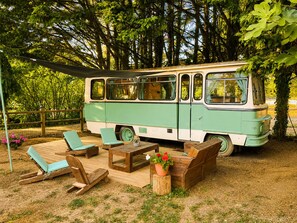 A vintage bus converted into a stationary living space with a deck, lounge chairs, and a table.