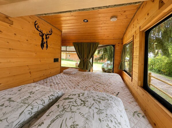 A cabin with wooden walls, a bed with floral bedding, a deer's head wall decal, and a window with a view of greenery.