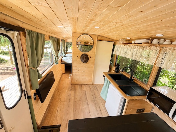 Interior compacto de autocaravana con kitchenette, un pequeño espacio de descanso y una ventana con cortinas.
