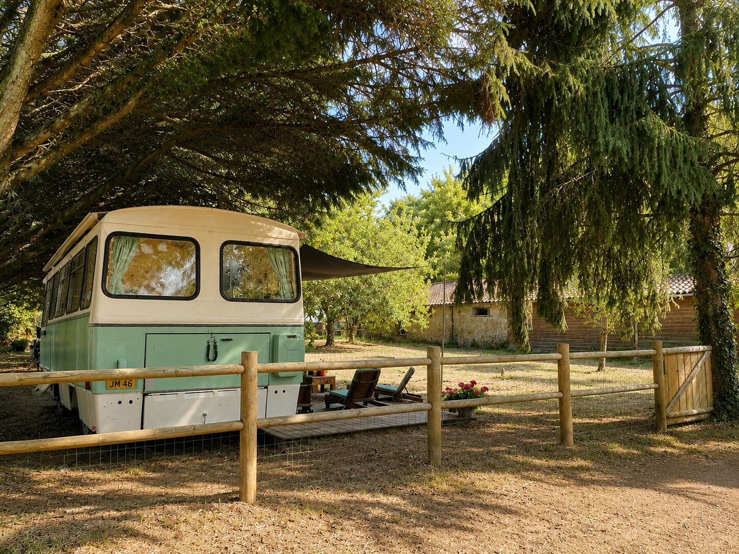 Un autobús antiguo estacionado en un área cercada por una valla de madera, rodeado de árboles y un edificio al fondo.