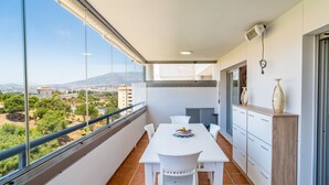 Un balcon avec une table et des chaises blanches, un vase et une vue sur le paysage urbain.