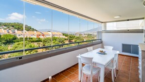 Un balcon avec une table et des chaises blanches, une vue sur des maisons et de la verdure, et un sol carrelé.