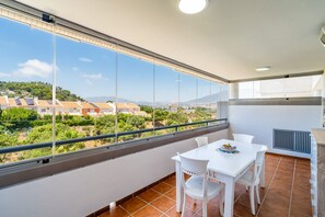 Ein Balkon mit weißem Tisch und Stühlen, Blick auf Häuser und Grünflächen, sowie ein Fliesenboden.