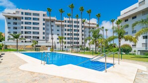 Une piscine aux bords recouverts de pierres, entourée d’une clôture, et des palmiers devant un bâtiment blanc doté de nombreuses fenêtres.