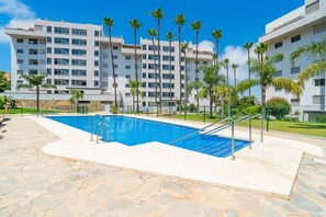 A swimming pool with stone tiles, surrounded by a fence, and palm trees in front of a white building with multiple windows. - Cubo's Vega Fuengirola Apartment & Parking / communal pool / terrace / free wifi (Fuengirola)