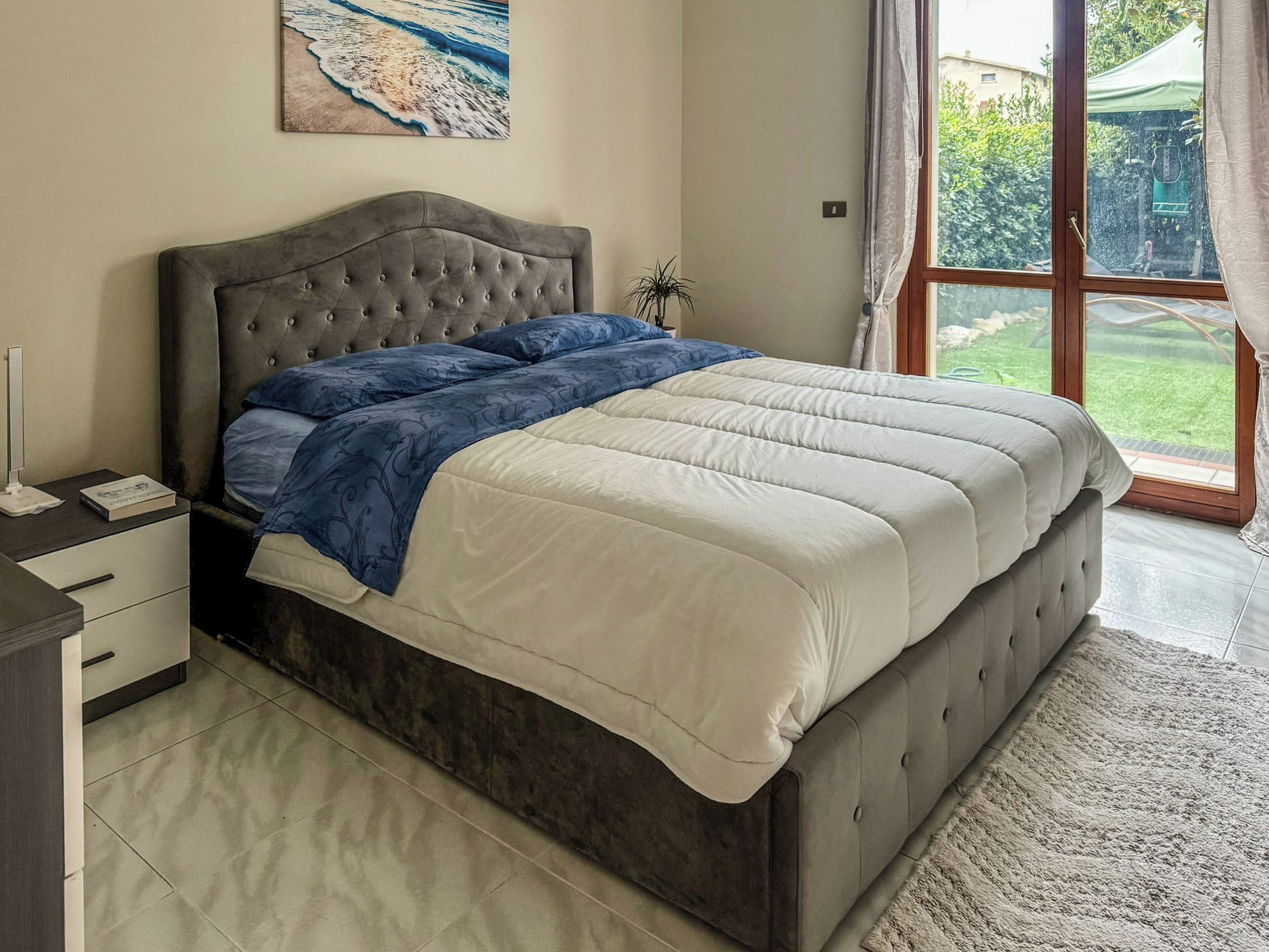 Une chambre avec un grand lit, une table de chevet, une commode, une fenêtre donnant sur un paysage verdoyant et un tableau accroché au mur.