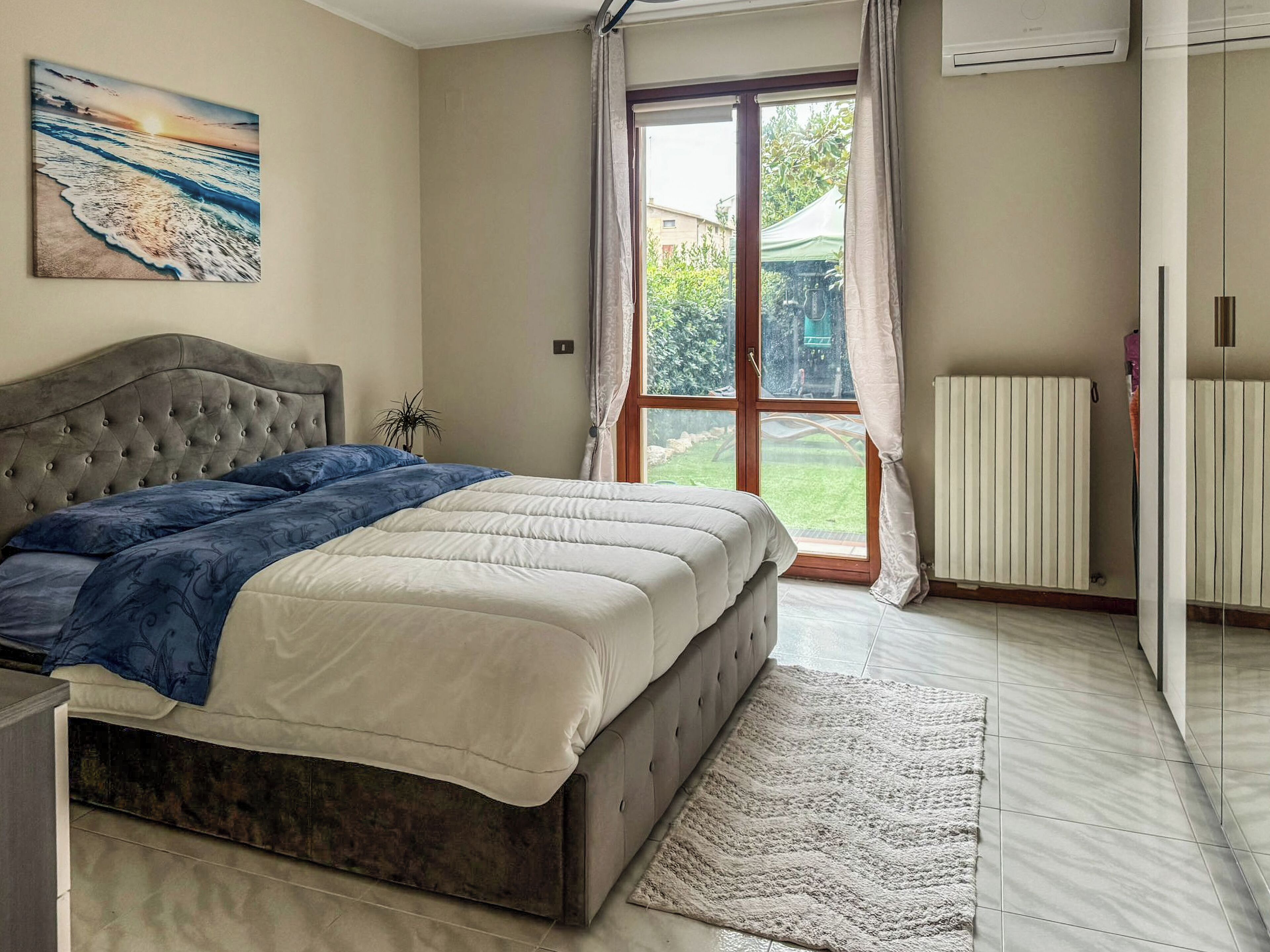 A bedroom with a bed, a radiator, a window with curtains, and a painting on the wall.
