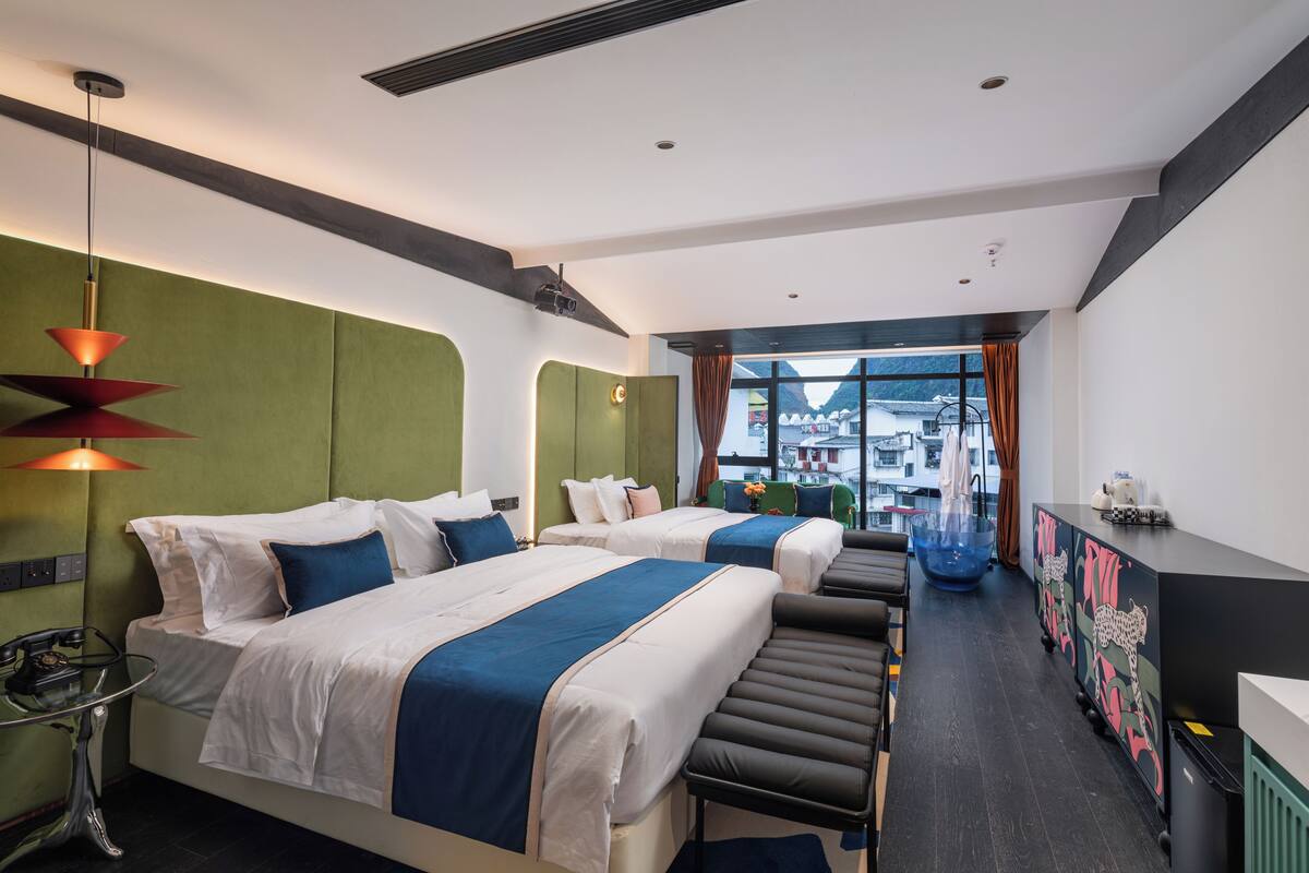 A hotel room with two beds, a desk, a chair, a sofa, a window with a view, and a decorative glass bowl.