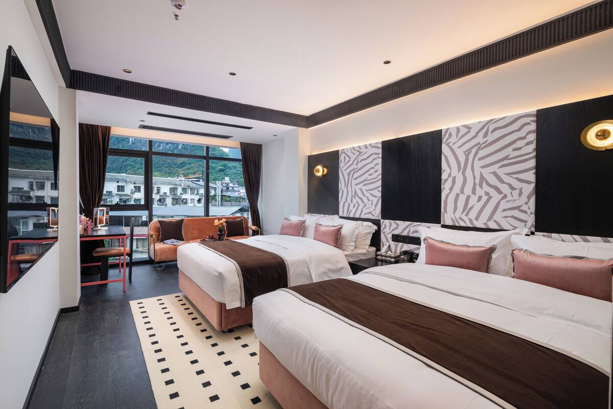 A modern hotel room with two beds, a desk, and a view of a mountainous landscape.