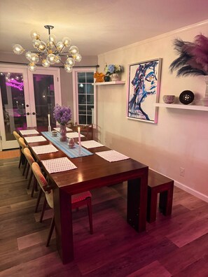 Ein Esszimmer mit einem langen Holztisch, Stühlen, einer Kandelaber, einem Gemälde und einer Tür zu einem weiteren Raum.