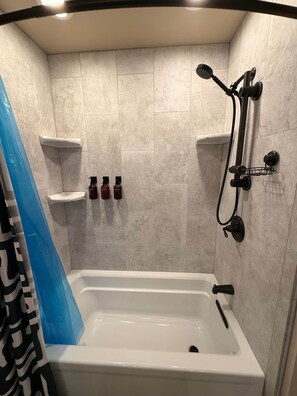 A bathroom with a bathtub, shower, and two shelves with bottles. - Brando King Room at Mountain Daddies B & B (Oakhurst)